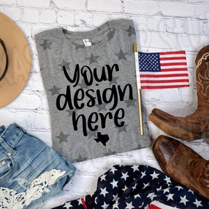 May include: A gray t-shirt with a white star pattern and the text "Your design here" in black. The shirt is laid flat on a white brick background with a pair of brown cowboy boots, a denim skirt, a brown hat, and an American flag.