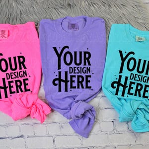 May include: Three t-shirts in pink, purple, and turquoise, each with the text "Your Design Here" in black. The shirts are knotted at the bottom and rest on a white brick surface.