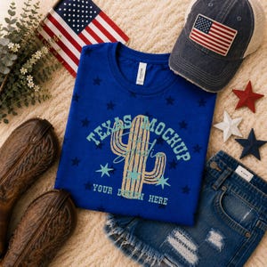 May include: A royal blue t-shirt featuring a gold cactus graphic, light blue stars, and the text "TEXAS MOCKUP STUDIO YOUR DESIGN HERE". The shirt is displayed alongside a distressed baseball cap with an American flag patch and brown cowboy boots.