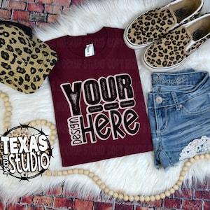 May include: A maroon t-shirt with the text "Design Your Here" in white and black lettering. The shirt is laying on a white faux fur surface with a leopard print hat, denim shorts with lace trim, and leopard print shoes.