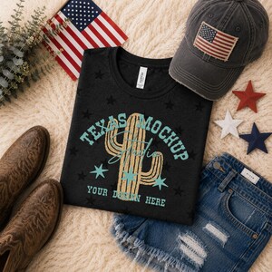 May include: A black t-shirt featuring a gold cactus graphic, teal stars, and the text "TEXAS MOCKUP STUDIO YOUR DESIGN HERE". The shirt is displayed with a charcoal baseball cap with an American flag patch, brown leather boots, and denim shorts.
