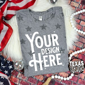 May include: A heather gray t-shirt with a star pattern and the text "Your Design Here" in white. The shirt is displayed on a white fluffy surface with an American flag and decorative beads.