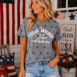May include: A grey short-sleeved t-shirt featuring a repeating pattern of dark grey stars. The center of the shirt displays white text that reads TEXAS MOCKUP Studio YOUR DESIGN HERE. The shirt is paired with light blue denim shorts with frayed hems.