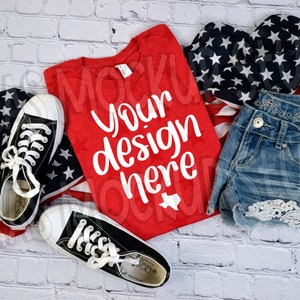 May include: Red t-shirt with the text "Your design here" in white. The shirt is laid flat on a white brick background with a pair of black sneakers, a pair of denim shorts with lace trim, and a blue and white American flag.