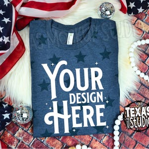 May include: A navy blue t-shirt with a star pattern and the text "YOUR DESIGN HERE" in white. The shirt is surrounded by patriotic decorations, including an American flag, disco balls, and a string of white beads. A Texas Mockup Studio logo is visible.