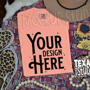 May include: A peach coloured t-shirt with the text "Your Design Here" in black lettering. The t-shirt is lying on a brick background with a pair of leopard print slip-on shoes, a straw hat, and a wooden bead necklace.
