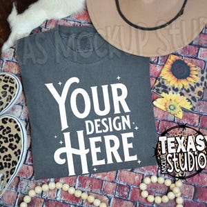 May include: A gray t-shirt with the text "YOUR DESIGN HERE" in white. The t-shirt is laying on a brick background with a brown hat, leopard print shoes, a sunflower coaster, and a wooden bead garland.