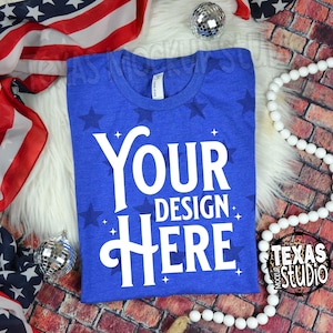 Könnte beinhalten: Ein königsblaues T-Shirt mit dem weißen Text "YOUR DESIGN HERE". Das Shirt hat einen Rundhalsausschnitt und ist mit dunkelblauen Sternen verziert. Der Hintergrund umfasst eine amerikanische Flagge, eine weiße, fellartige Oberfläche und dekorative Perlen.