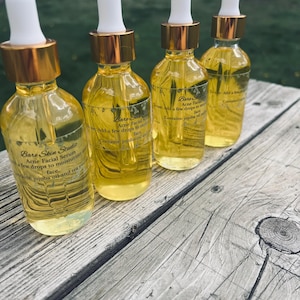 Może przedstawiać: Cztery szklane butelki serum do twarzy Bare Skin Studio Acne Facial z białymi zakraplaczami i złotymi nakrętkami. Butelki są wypełnione ż&oacute;łtym płynem i ułożone na zwietrzałej drewnianej powierzchni. Tekst na etykiecie zawiera "jojoba oil and tea tree".