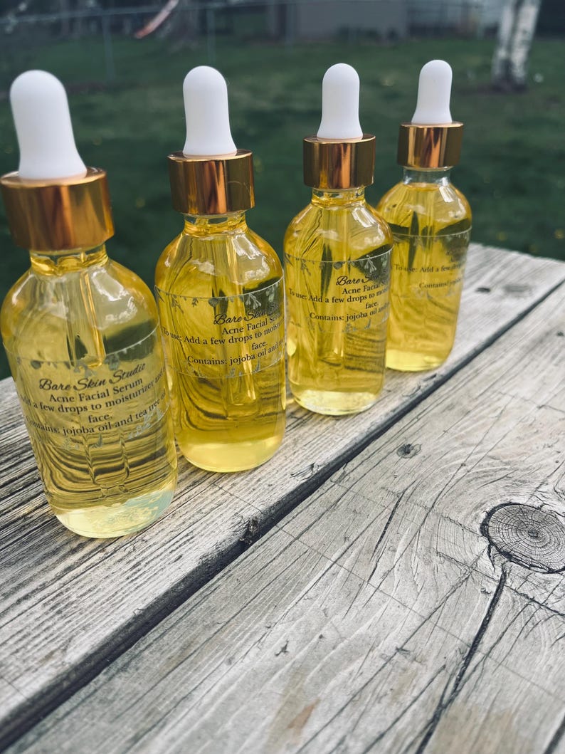 Może przedstawiać: Cztery szklane butelki serum do twarzy Bare Skin Studio Acne Facial z białymi zakraplaczami i złotymi nakrętkami. Butelki są wypełnione ż&oacute;łtym płynem i ułożone na zwietrzałej drewnianej powierzchni. Tekst na etykiecie zawiera "jojoba oil and tea tree".