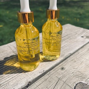 Może przedstawiać: Dwie szklane butelki serum do twarzy Bare Skin State Acne z białymi zakraplaczami i złotymi nakrętkami. Butelki zawierają ż&oacute;łty płyn i są oznaczone informacjami o produkcie. Na drewnianej powierzchni.