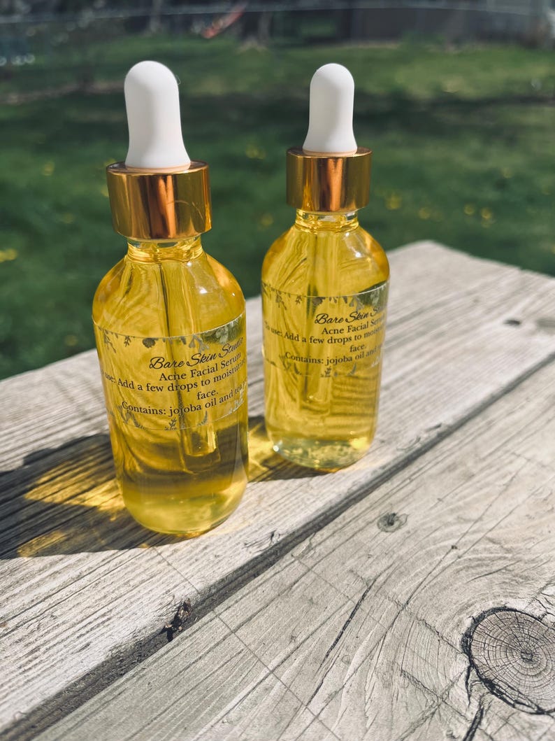 Może przedstawiać: Dwie szklane butelki serum do twarzy Bare Skin State Acne z białymi zakraplaczami i złotymi nakrętkami. Butelki zawierają ż&oacute;łty płyn i są oznaczone informacjami o produkcie. Na drewnianej powierzchni.