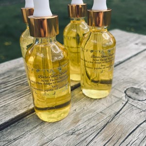 Może przedstawiać: Cztery szklane butelki serum do twarzy przeciwtrądzikowego Bare Skin Studio z białymi zakraplaczami i złotymi kołnierzami. Butelki są wypełnione ż&oacute;łtym płynem i stoją na drewnianej powierzchni. Etykieta zawiera informację o oleju jojoba i herbacie.