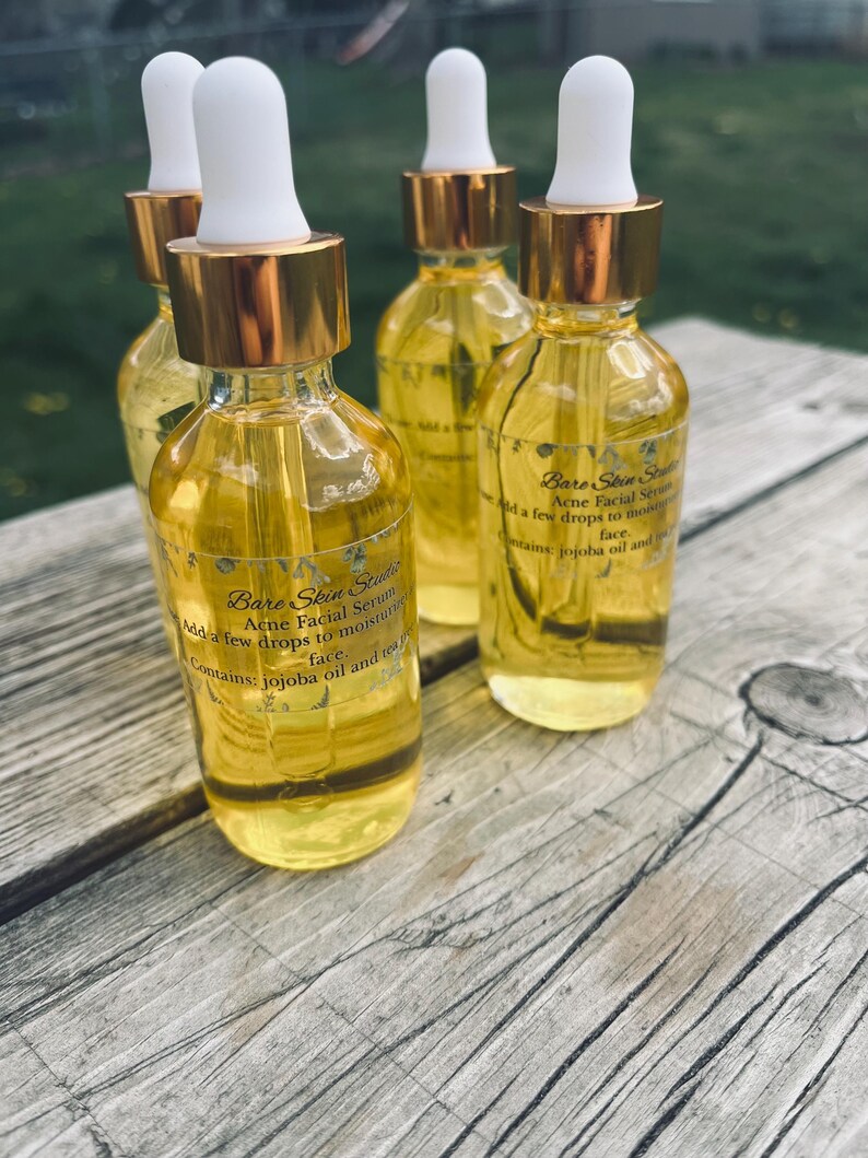 Może przedstawiać: Cztery szklane butelki serum do twarzy przeciwtrądzikowego Bare Skin Studio z białymi zakraplaczami i złotymi kołnierzami. Butelki są wypełnione ż&oacute;łtym płynem i stoją na drewnianej powierzchni. Etykieta zawiera informację o oleju jojoba i herbacie.