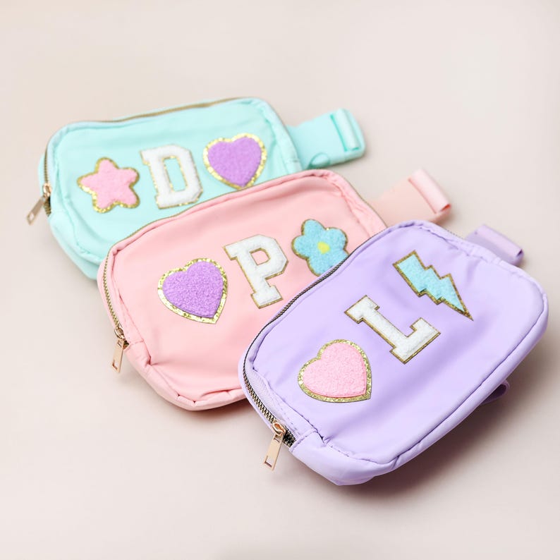 May include: Three pastel-coloured fanny packs in mint, pink, and lavender. Each pack has a zip closure, adjustable strap, and decorative patches including letters, hearts, stars, flowers, and a lightning bolt.
