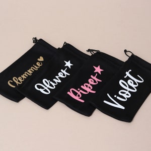 May include: Four black drawstring bags with different names printed on them in white, gold, and pink lettering. The names are "Clemmie", "Oliver", "Piper", and "Violet".