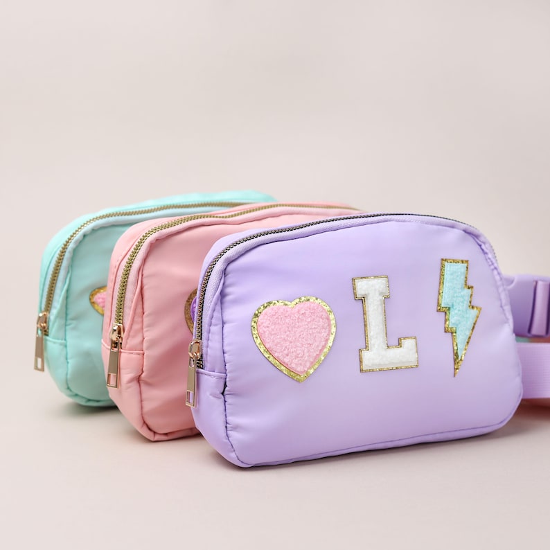 May include: Three pastel-coloured fanny packs in mint green, pink, and lavender. The lavender pack has a heart, the letter "L", and a lightning bolt patch. Each pack has a gold zip and a matching adjustable strap.