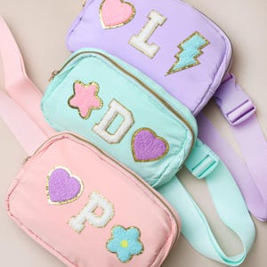 May include: Three pastel-coloured fanny packs in pink, mint green, and lavender. Each pack features a different letter and decorative patches, including hearts, stars, and a lightning bolt. The packs have adjustable straps and gold zips.