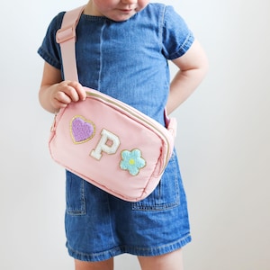 May include: A pink crossbody bag with a gold zip and strap. The bag is decorated with a purple heart, a white letter "P", and a blue and green flower. The bag is worn by a person.