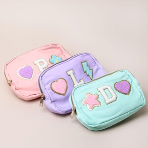 May include: Three small, zippered cosmetic bags in pastel shades: pink, lavender, and mint green. Each bag features a letter, a heart, and other decorative patches. The bags have gold-coloured zips and are arranged on a neutral background.