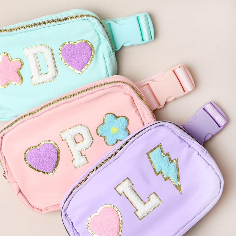 May include: Three pastel-coloured fanny packs in mint, pink, and lavender. Each pack has a gold zip and decorative patches, including letters, hearts, stars, flowers, and a lightning bolt. The packs have adjustable straps and plastic buckles.