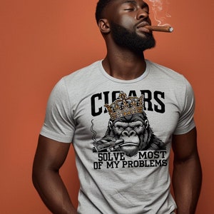 以下が含まれることがあります： 王冠をかぶって葉巻を吸っているゴリラのグラフィックがプリントされたグレーのTシャツ。ゴリラの真下に「CIGARS SOLVE MOST OF MY PROBLEMS」というテキストがプリントされています。