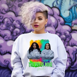 May include: White sweatshirt featuring a graphic of two women with long black hair, set against a colorful background. The text "THE BOY IS MINE Tour" is printed below the image in green. The model has purple and white hair.