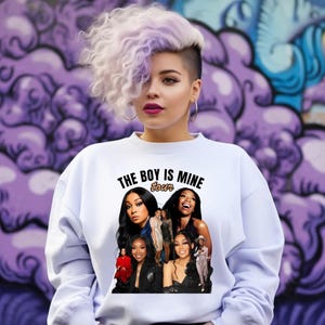 Pode incluir: Camisola branca com o texto "THE BOY IS MINE tour" em preto. A camisola apresenta uma colagem de imagens de várias mulheres. O fundo é uma parede de grafite roxa.