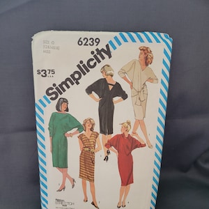 May include: A vintage Simplicity sewing pattern featuring illustrations of dresses. The pattern cover is white with a blue and white striped border and the Simplicity logo. The pattern number is 6239, and the size is O (12, 14, & 16).