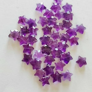 May include: A collection of small, star-shaped amethyst gemstones. The faceted stones display varying shades of purple, from light lavender to deep violet. The stars are scattered against a white background.