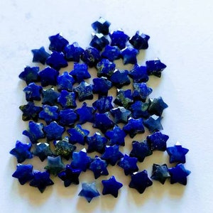 May include: A collection of small, star-shaped lapis lazuli gemstones. The gemstones are a deep blue color with flecks of gold and are scattered across a white surface. These faceted stars are ideal for jewelry making or craft projects.