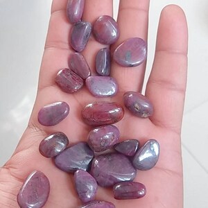 May include: A collection of polished, oval-shaped gemstones in varying shades of red and purple. The stones are smooth and reflective, with a natural, organic appearance. The gemstones are held in a hand.