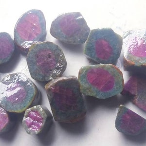 Puede incluir: Una colección de piedras preciosas de rubí en bruto. Cada piedra tiene un centro rosa vibrante rodeado por una capa exterior azul-verdosa. Las piedras varían en tamaño y forma, con una textura rugosa y natural.