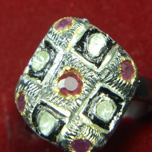 Peut inclure: Bague argentée avec un motif carré texturé. La bague présente des carrés clairs et noirs alternés, chacun contenant une petite pierre précieuse ronde. De petites pierres rouges sont également serties dans le motif.
