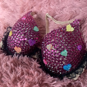 May include: A pink bra with a black lace trim, covered in rhinestones and colorful heart-shaped embellishments.