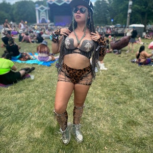 May include: A woman wearing a silver and black bra with fringe, leopard print shorts, and silver platform boots. She is wearing a black baseball cap with the word "OPEN" on it. She is standing in a grassy field with a crowd of people in the background.