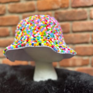 May include: A colorful bucket hat covered in small, vibrant pom-poms. The hat has a wide brim and a light-colored interior. The hat is displayed on a white mushroom-shaped stand against a brick background.
