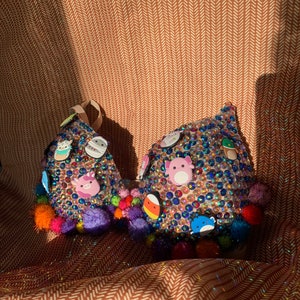 May include: A sparkly pink bra decorated with colorful pom-poms and various cartoon character charms. The bra is covered in rhinestones and has a light pink base.
