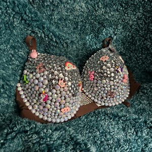 Puede incluir: Un sujetador marrón adornado con pedrería iridiscente y pequeños abalorios de colores. Los abalorios incluyen un arcoíris, un unicornio y varios otros diseños bonitos. El sujetador tiene tirantes y copas marrones.