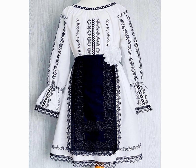 May include: Traditional white and navy blue dress with black embroidery. The dress has long sleeves with flared cuffs, a floral skirt panel, and a decorative flower. The neckline and hem are trimmed with black detailing.
