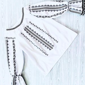 May include: White blouse featuring black embroidery. The design includes detailed patterns along the neckline and sleeves. The cuffs have a ruffled trim. The garment is made from a light fabric.