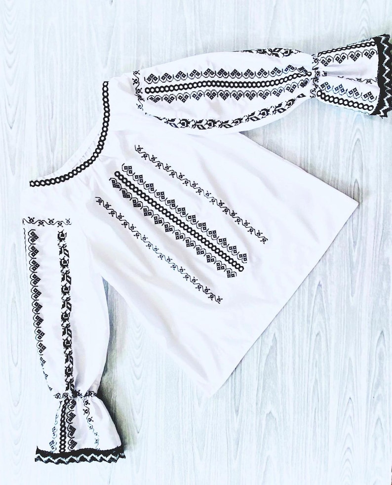 May include: White blouse featuring black embroidery. The design includes detailed patterns along the neckline and sleeves. The cuffs have a ruffled trim. The garment is made from a light fabric.