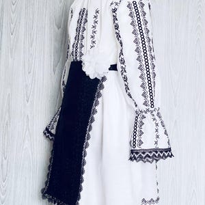 May include: White dress with black embroidery and lace edging. The dress has long sleeves with flared cuffs, a black sash, and a white flower. It is designed with traditional folk elements.