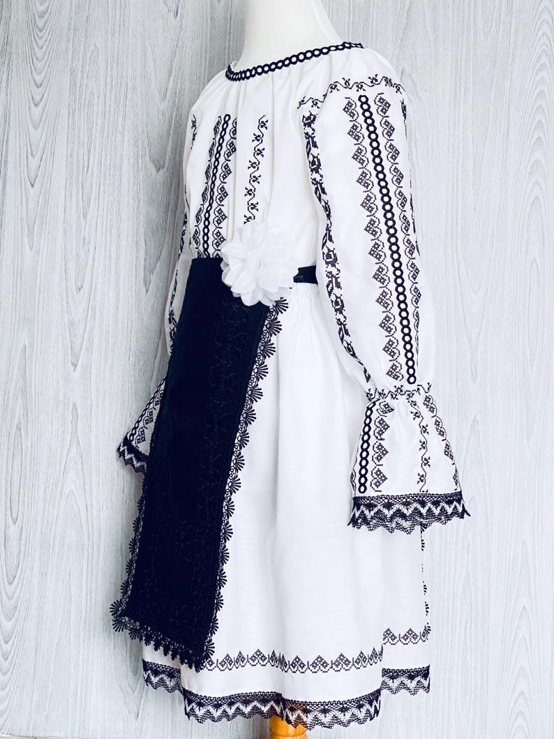 May include: White dress with black embroidery and lace edging. The dress has long sleeves with flared cuffs, a black sash, and a white flower. It is designed with traditional folk elements.