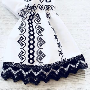 May include: White fabric sleeve with black embroidered floral patterns and a black lace trim. The sleeve features a gathered detail and a flared shape, showcasing traditional embroidery.