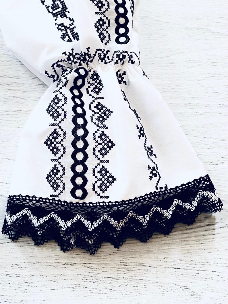 May include: White fabric sleeve with black embroidered floral patterns and a black lace trim. The sleeve features a gathered detail and a flared shape, showcasing traditional embroidery.