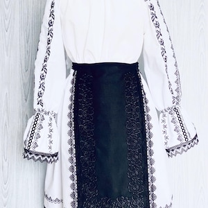 May include: A white dress with long sleeves and detailed black embroidery. The dress has a black belt and a black panel with floral patterns. The sleeves have flared cuffs with matching embroidery, and the neckline is trimmed with black.