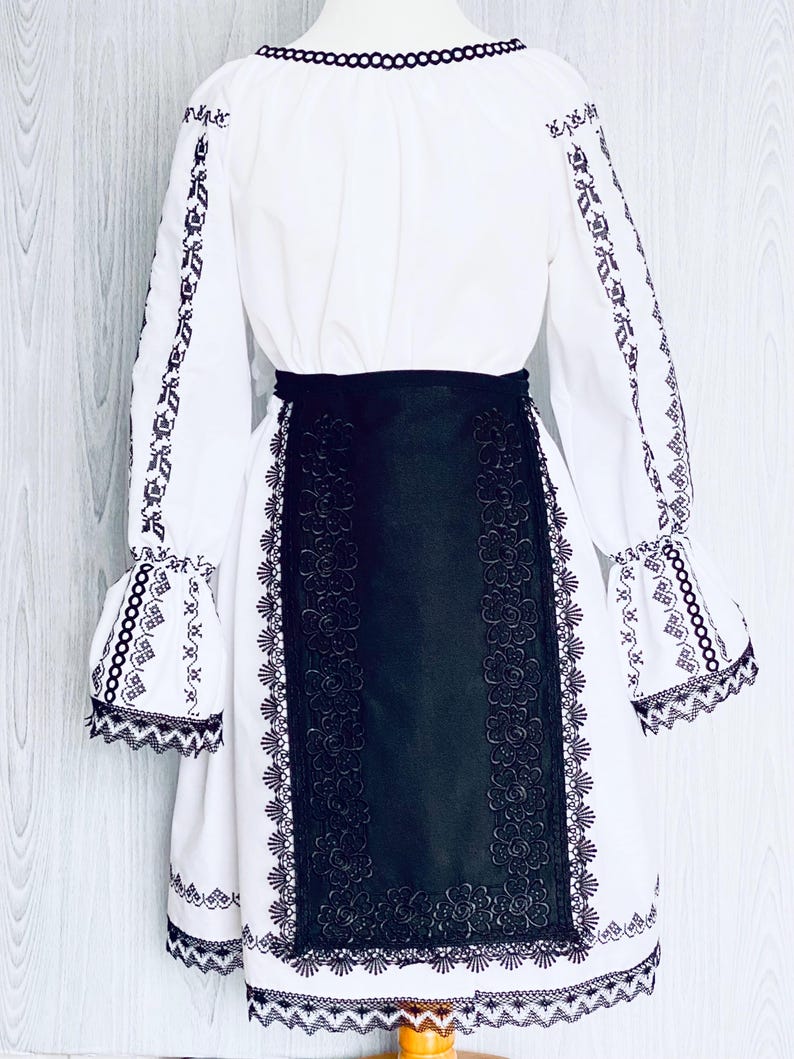 May include: A white dress with long sleeves and detailed black embroidery. The dress has a black belt and a black panel with floral patterns. The sleeves have flared cuffs with matching embroidery, and the neckline is trimmed with black.