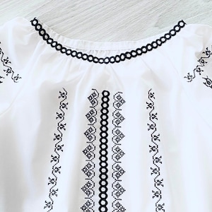 May include: White blouse with black embroidered details. The neckline and sleeves are trimmed with a black scalloped design. The front of the blouse features vertical and horizontal embroidered patterns. The embroidery is in a geometric and floral style.