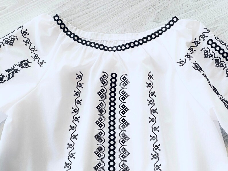 May include: White blouse with black embroidered details. The neckline and sleeves are trimmed with a black scalloped design. The front of the blouse features vertical and horizontal embroidered patterns. The embroidery is in a geometric and floral style.